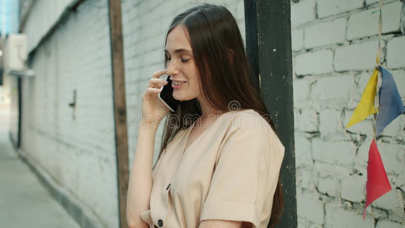 Vue de côté de belle jeune dame bavardant sur le téléphone portable et souriant debout à l'extérieur près de mur de brique banque de vidéos