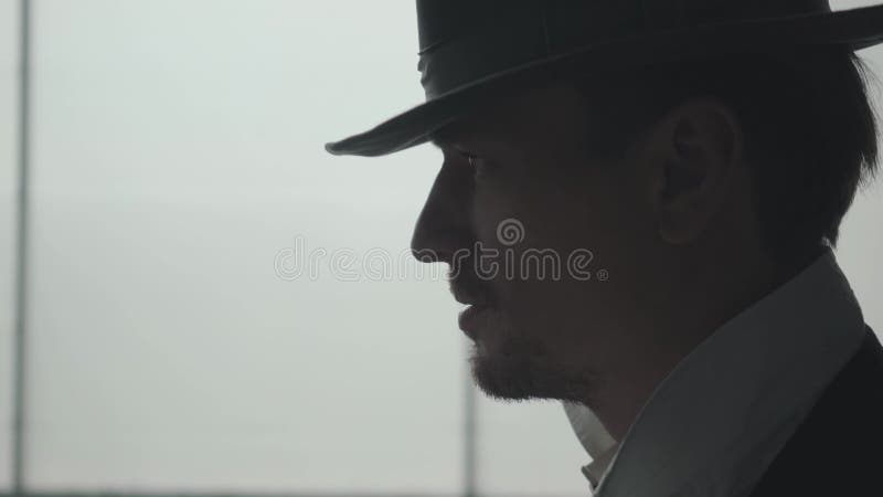 Vue de côté de l'homme sûr bel enlevant le chapeau regardant en avant avec le regard fixe menaçant devant la grande fenêtre La t? banque de vidéos