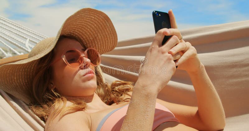 Vue de côté de la femme caucasienne à l'aide du téléphone portable dans un hamac sur la plage 4k banque de vidéos