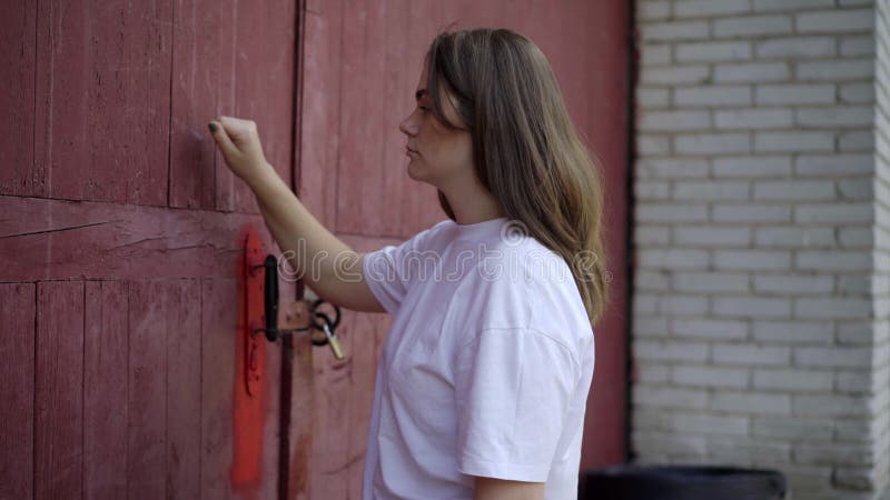 Vue de côté de la jeune belle caucasienne brune femme frapper dans grande porte rouge à l extérieur. portrait de charme sérieux clips vidéos