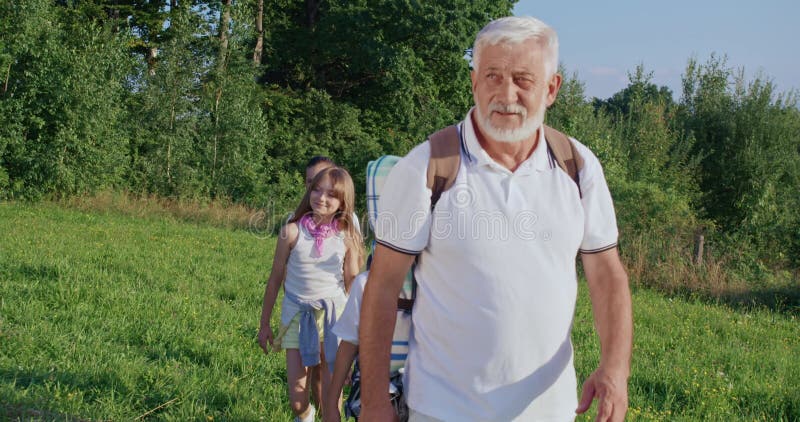 Vue de devant du vieil homme avec un sac à dos groupe d'enfants de chef de file suivant derrière lui des enfants souriant parler. banque de vidéos