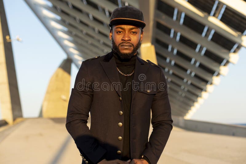 Vue de face du jeune homme élégant noir se tenant dehors dans un jour ensoleillé tout en regardant la caméra photographie stock