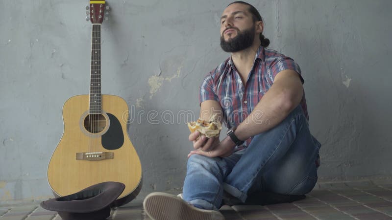Vue de face de tir large du jeune homme frustrant assis dans le passage souterrain avec la guitare et la consommation Portrait de banque de vidéos