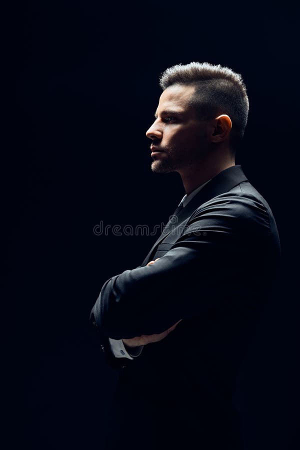 Vue de profil d'homme sûr dans le costume noir avec des bras croisés sur le fond foncé photos libres de droits Vue de profil d'homme sûr dans le costume noir avec des bras croisés sur le fond foncé photos libres de droits