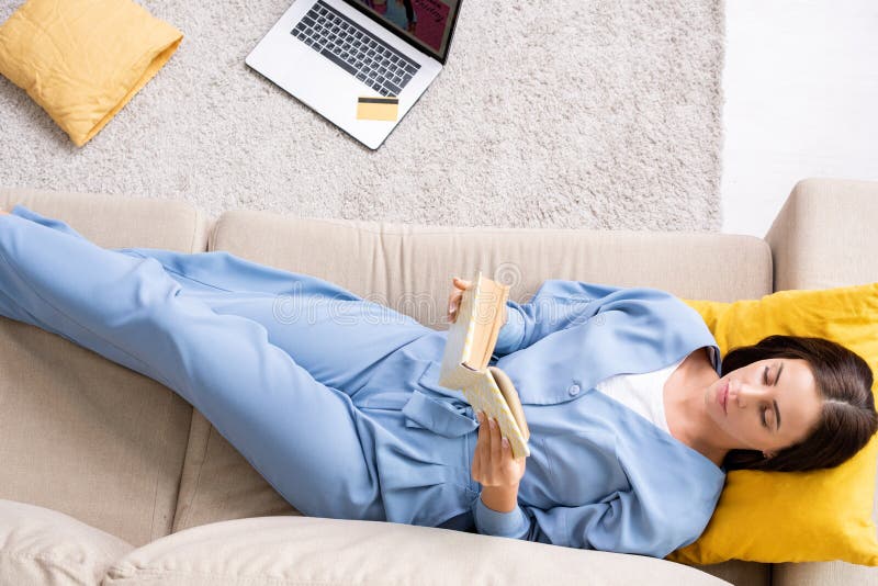 Vue du haut de la jeune femme reposante en pyjama bleu lecture livre sur le canapé à la maison photographie stock libre de droits