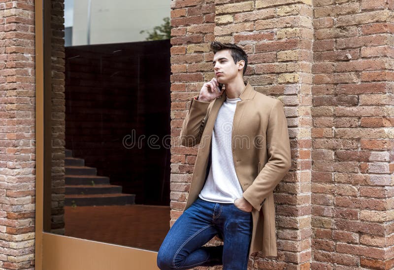 Vue frontale d'un jeune homme à la mode portant des vêtements en denim penchés sur un mur en utilisant un téléphone portable à l'e images libres de droits