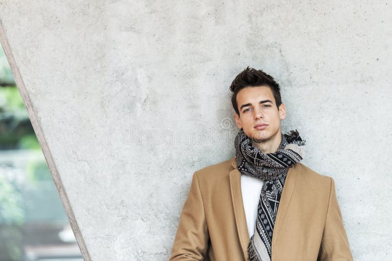 Vue frontale d'un jeune homme élégant vêtu d'un manteau et d'une écharpe penché sur un mur en regardant la caméra images stock
