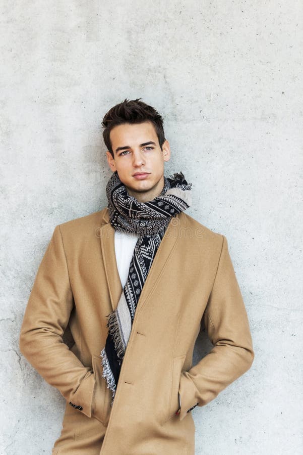 Vue frontale d'un jeune homme élégant vêtu d'un manteau et d'une écharpe sur un mur tout en regardant loin image libre de droits