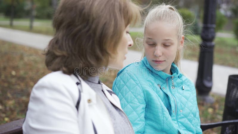Vue latérale de deux femmes caucasiennes d'âges différents assises dans le parc ensoleillé et parlant Adolescent blond irrité banque de vidéos
