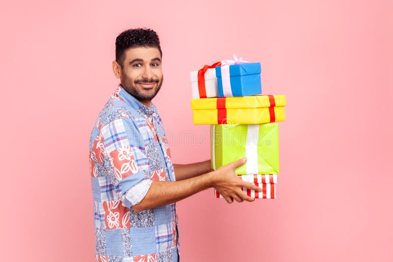 Vue latérale de l'homme barbu tenant pile de cadeaux dans les mains en regardant la caméra avec un sourire agréable célébrant les photos libres de droits