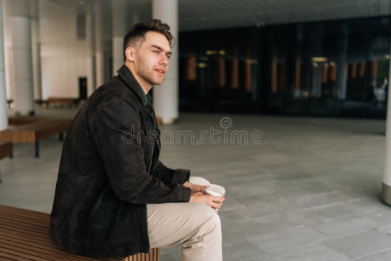 Vue latérale du jeune homme pensif assis sur un banc en bois dans un environnement urbain contemporain tenant deux tasses de café image libre de droits