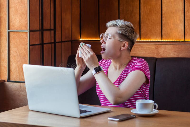 Vue latérale portrait de la maladie jeune femme d'affaires aux cheveux blonds courts en t-shirt rose et lunettes assis au café et photographie stock