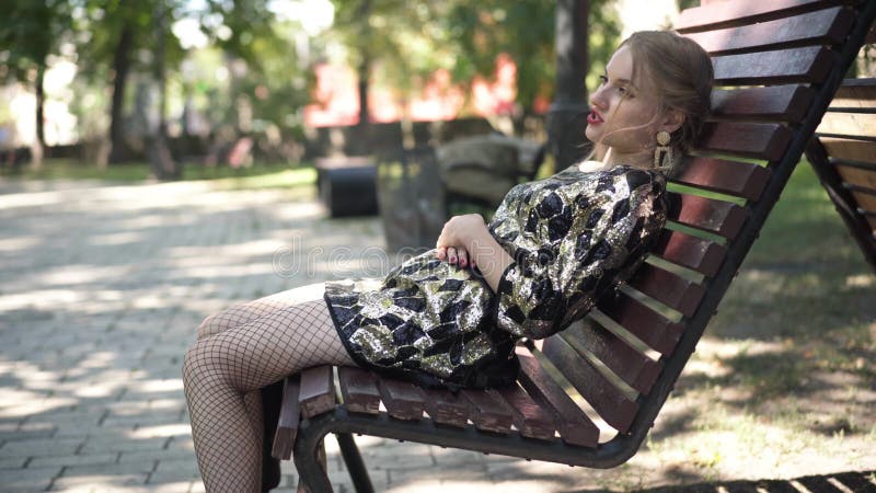 Vue latérale une jeune femme caucasienne insouciante en tenue de fête assise sur un banc dans le parc qui parle de bâillonnement. banque de vidéos