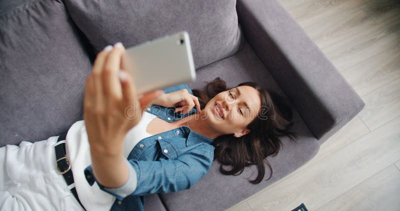 Vue supérieure de la belle jeune dame prenant le selfie avec le smartphone se trouvant sur le sofa banque de vidéos