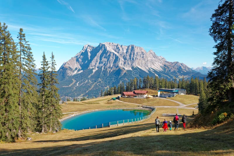 Vue sur la montagne escarpée Zugspitze par une journée ensoleillée avec des touristes se promenant vers un étang bleu au pied des photo stock Vue sur la montagne escarpée Zugspitze par une journée ensoleillée avec des touristes se promenant vers un étang bleu au pied des photo stock