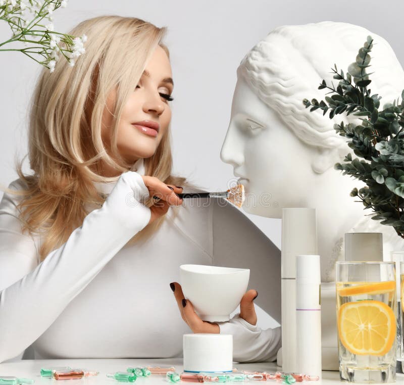 Vuxen blond kvinna i pullovrar vid bord med kosmetiska produkter innebär att man använder statushuvudets ansiktsmask med pensel royaltyfri foto
