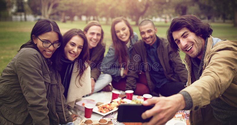 Vänner som är glada och själviska i parken för picknikbindning och sociala medier på helgen med snacks på sommaren. grupp av perso royaltyfri bild