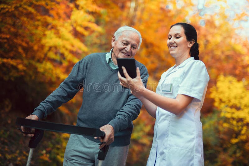 Vårda och den rörelsehindrade höga patienten i fotgängaren som använder den digitala minnestavlan arkivfoto Vårda och den rörelsehindrade höga patienten i fotgängaren som använder den digitala minnestavlan arkivfoto