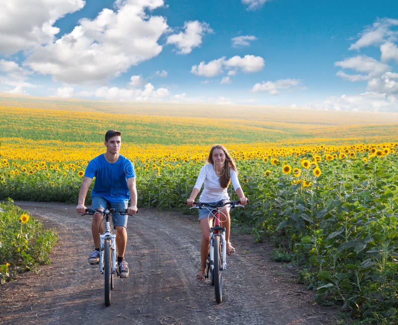 Vélo de l'adolescence d'équitation de couples dans le domaine de tournesol photographie stock libre de droits Vélo de l'adolescence d'équitation de couples dans le domaine de tournesol photographie stock libre de droits