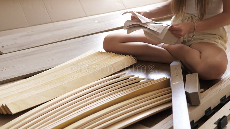 Vídeo de cierre de la joven está leyendo instrucción en las listas de papel para montar una cama de madera sentada en el piso de l