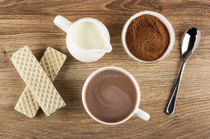 Wafers with porous chocolate, jug of milk, cocoa with sugar in bowl, cup of cocoa with milk, spoon on table. Top view