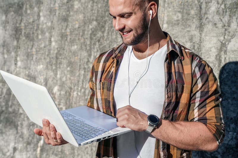 Waist up portrait of young good looking bold bearded hipster standing outdoors against grey modern loft wall with laptop and stock image Waist up portrait of young good looking bold bearded hipster standing outdoors against grey modern loft wall with laptop and stock image