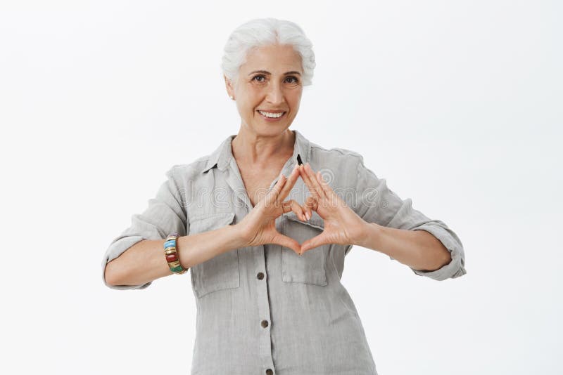 Waist-up shot of charming caring granny with grey hair in shirt showing heart sign over chest and smiling broadly loving stock image Waist-up shot of charming caring granny with grey hair in shirt showing heart sign over chest and smiling broadly loving stock image
