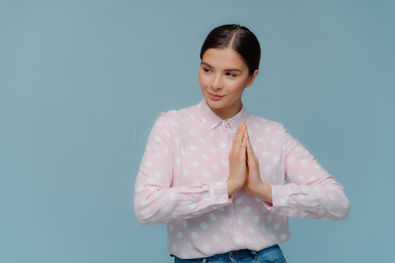 Waist up shot of lovely woman presses palms together, prays indoor, focused away, wears polka dot shirt, has serious thoughtful stock photo Waist up shot of lovely woman presses palms together, prays indoor, focused away, wears polka dot shirt, has serious thoughtful stock photo