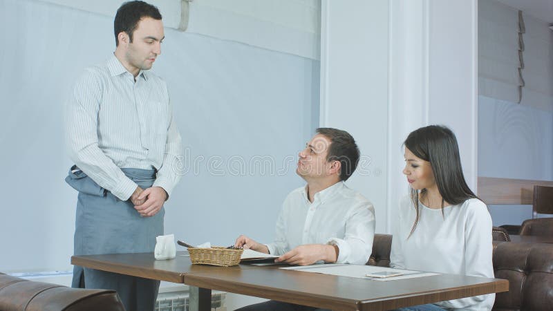 Waiter bringing menu to young couple sitting at the table in a restaurant