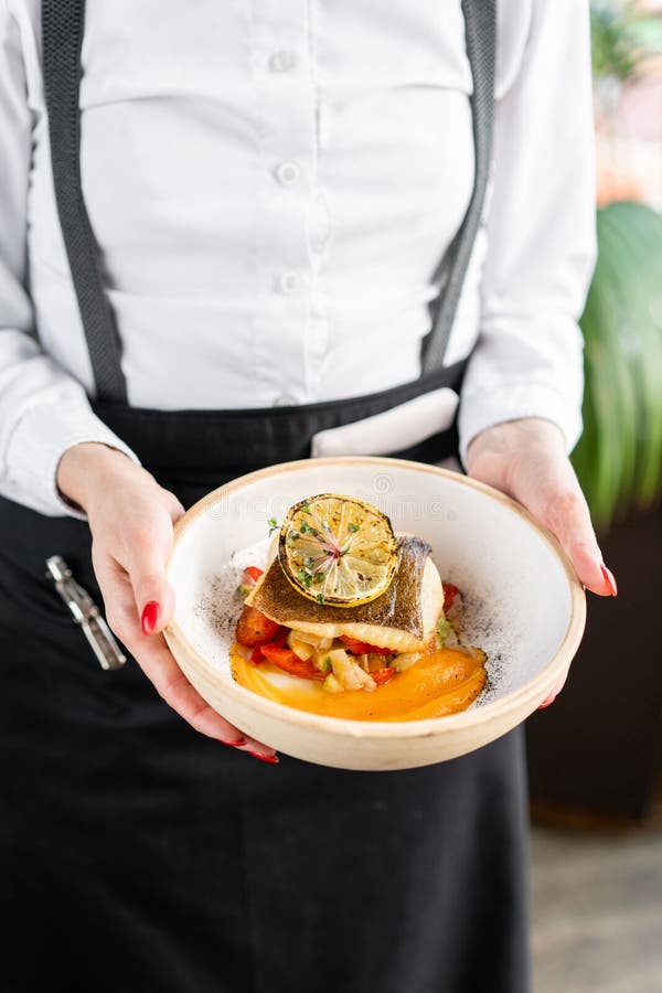 The waiter is holding a plate Roasted pike perch or cod fish with baked vegetables. Dish decorated with a slice of lemon stock photo