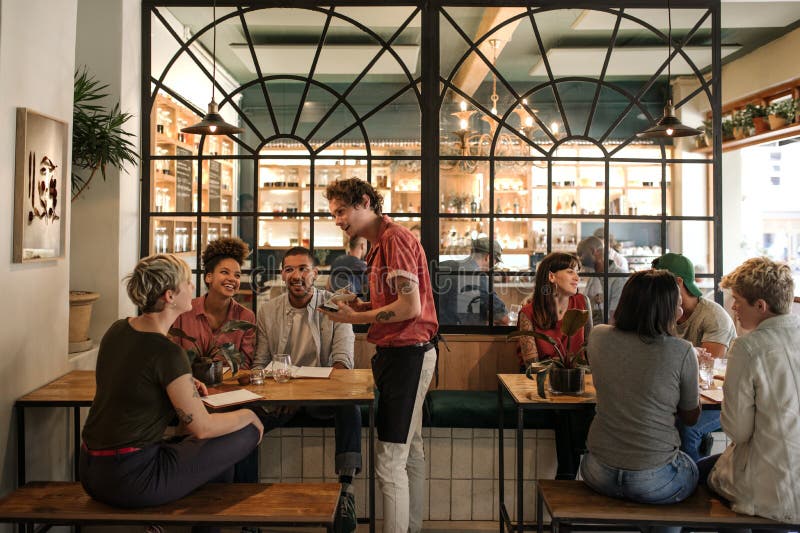 Waiter taking orders from customers sitting in a bistro. Smiling young waiter taking orders from a diverse group of customers sitting together at a restaurant table
