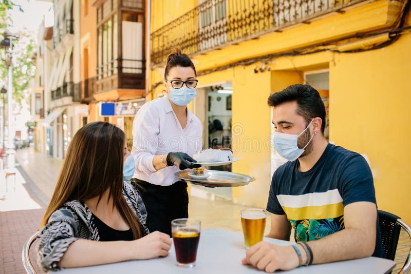 Waitress from a bar in Spain serves food and drinks to customers on her terrace. Everyone wears a mask to protect themselves from. Contagion. Phase one of the stock photos Waitress from a bar in Spain serves food and drinks to customers on her terrace. Everyone wears a mask to protect themselves from. Contagion. Phase one of the stock photos