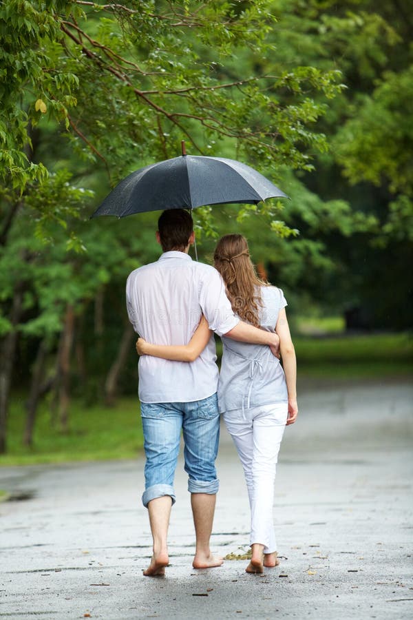 Rear view of a couple walking in the rain under umbrella and hugging.