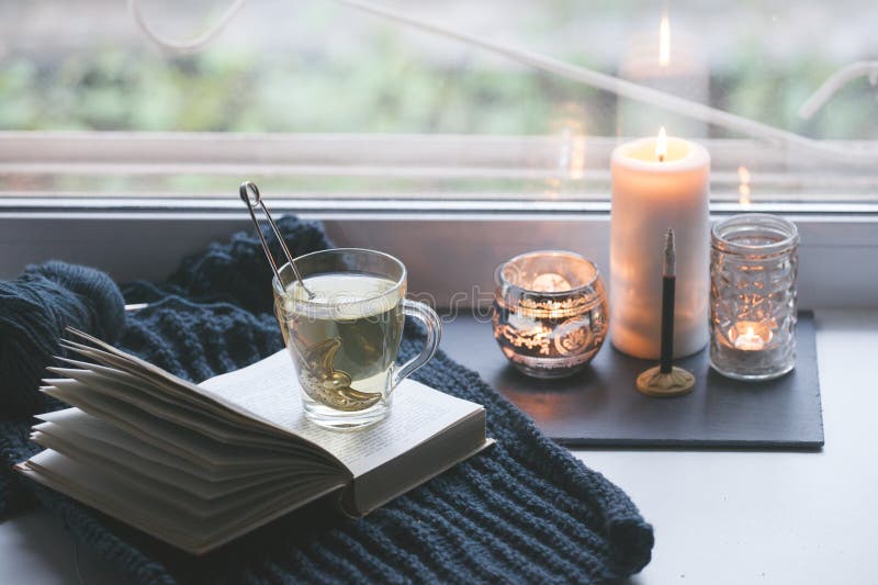Warm and comfy autumn concept. Book, cup of tea and candlestick on window sill in house. Reading and relaxing in cold weather at