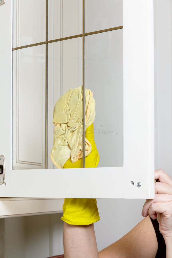 Female hands in yellow rubber gloves wipe the glass door of the kitchen cabinet with a rag. Female hands in yellow rubber gloves wipe the glass door of the kitchen cabinet with a rag
