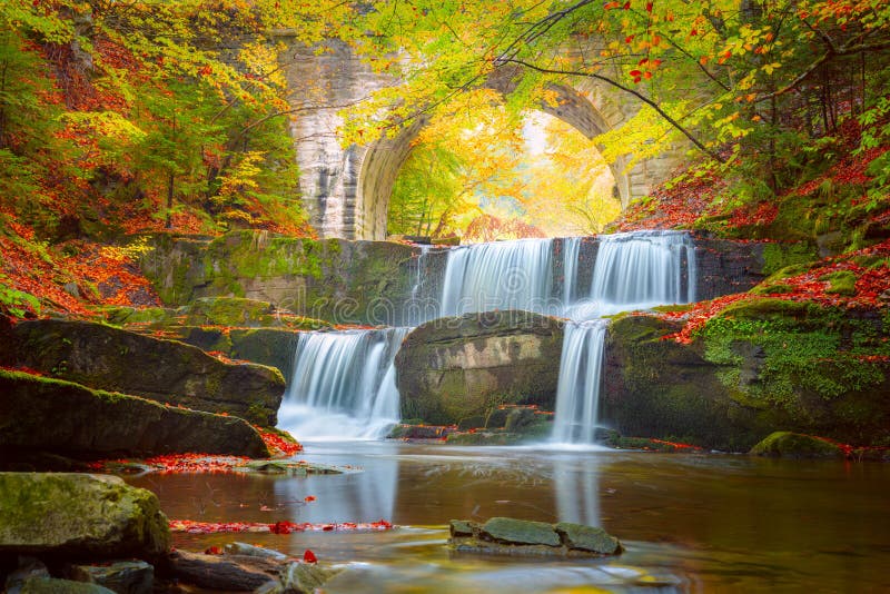 Wasserfall der Herbstnaturlandschaft- Fluss im bunten Herbstwald mit alter Brücke stockfotografie
