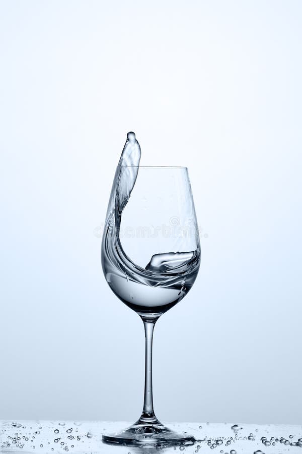 Water splashing from wineglass with cleaner water while standing on the glass with droplets against light background.