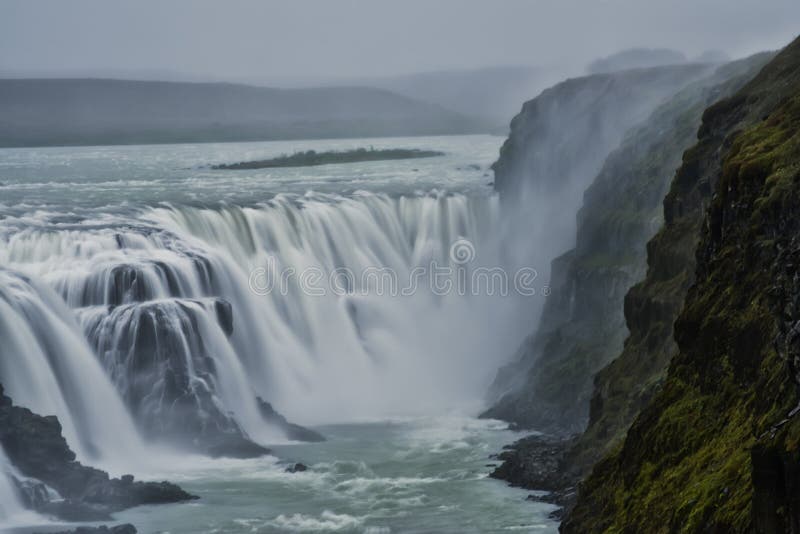 A number of steep cliffs on the right and a huge waterfall. Gloomy, severe weather, gloomy sky. Everything is shrouded in smoke from the waterfall. Nature and sights of Iceland. A number of steep cliffs on the right and a huge waterfall. Gloomy, severe weather, gloomy sky. Everything is shrouded in smoke from the waterfall. Nature and sights of Iceland.