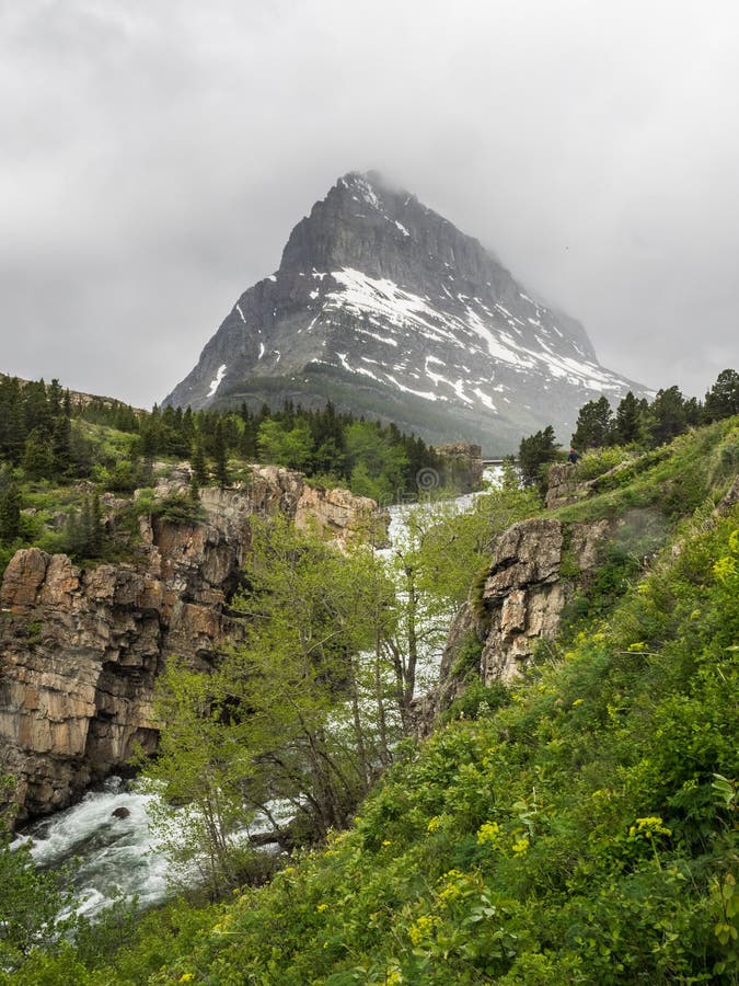 Waterfalls and snow capped mountain. Water tumbles through a rocky canyon with a snow-capped mountain on a cloudy day at Many Glacier in Glacier National Park stock photo Waterfalls and snow capped mountain. Water tumbles through a rocky canyon with a snow-capped mountain on a cloudy day at Many Glacier in Glacier National Park stock photo