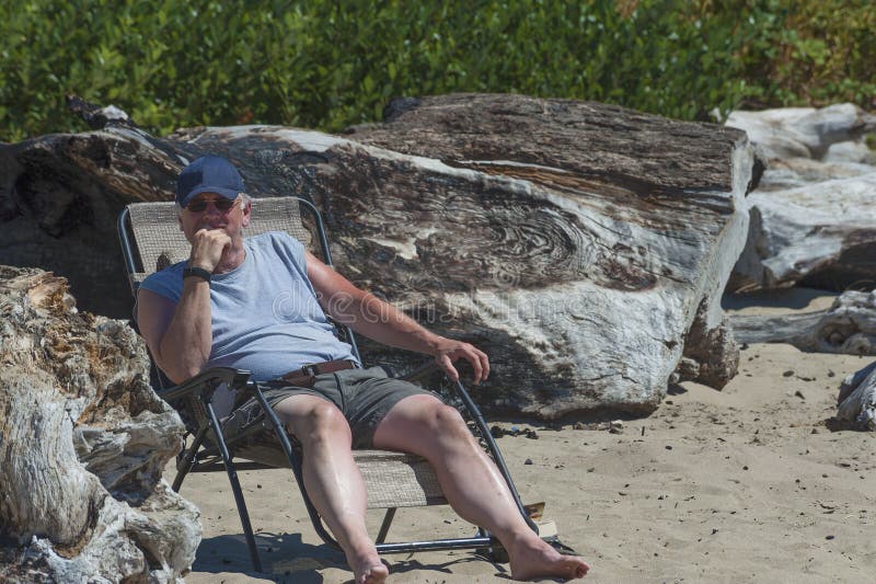 Wearing a ball cap and sunglasses an elderly man lounges on a folding chaise near large driftwood on beach at Siletz Bay Park in Lincoln City, Oregon. Wearing a ball cap and sunglasses an elderly man lounges on a folding chaise near large driftwood on beach at Siletz Bay Park in Lincoln City, Oregon