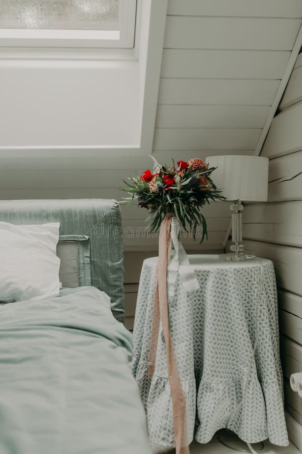 A wedding bouquet of red flowers decorated with a ribbon stands on the bedside table in the room.