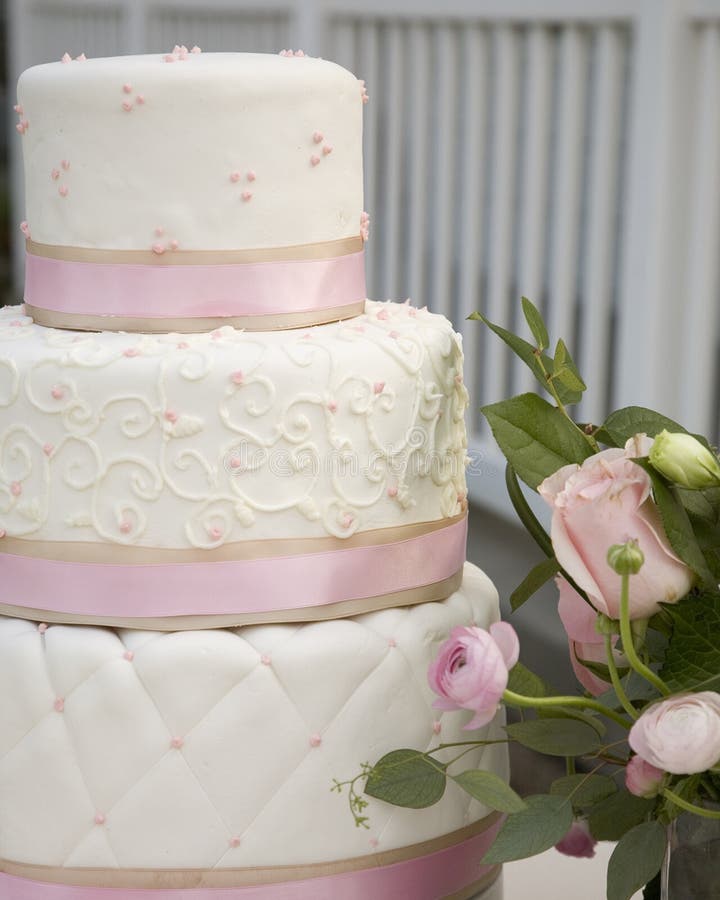 Close up di tiered bianco laminati fondente torta nuziale, strati avvolti con rosa e khaki nastro decorato con motivo trapuntato, turbinii e puntini accanto a una rosa fresca e floreale.