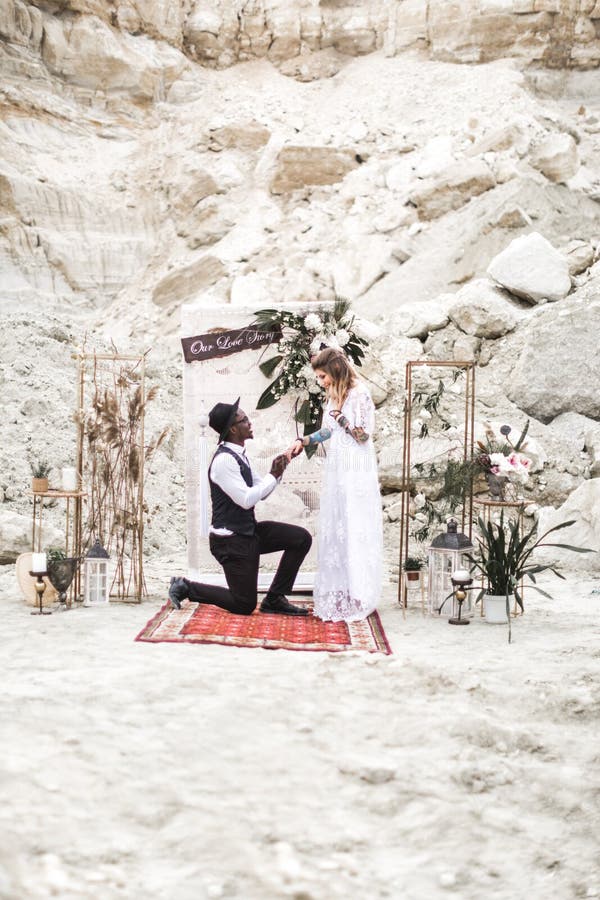 Wedding couple dressed in boho style are staying before the wedding arch, holding hands. African groom is standing on royalty free stock photo Wedding couple dressed in boho style are staying before the wedding arch, holding hands. African groom is standing on royalty free stock photo