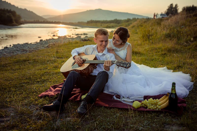 Wedding picnic composition on the river bank during the sunset. Smiling groom is playing the guitar while bride is