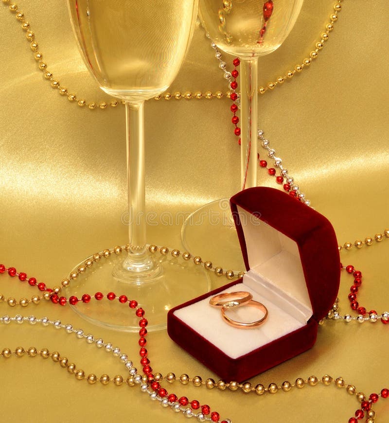 Wedding rings and glasses with sparkling wine on a golden background. A composition Valentine's Day: Satin drapery, decorated with beads, which is velvet red box with wedding rings and there are two glasses of sparkling wine, clouse up