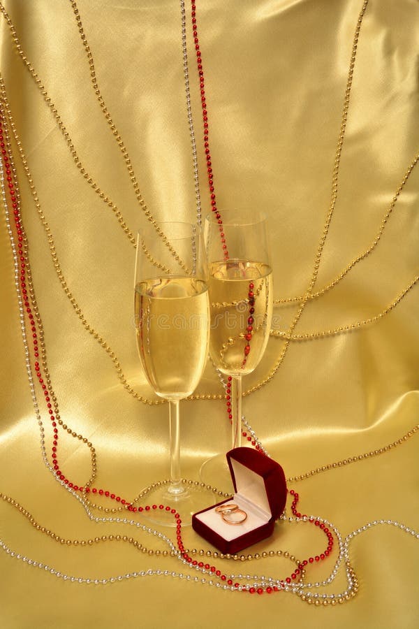 Wedding rings and glasses with sparkling wine on a golden background. A composition Valentine's Day: Satin drapery, decorated with beads, which is velvet red box with wedding rings and there are two glasses of sparkling wine