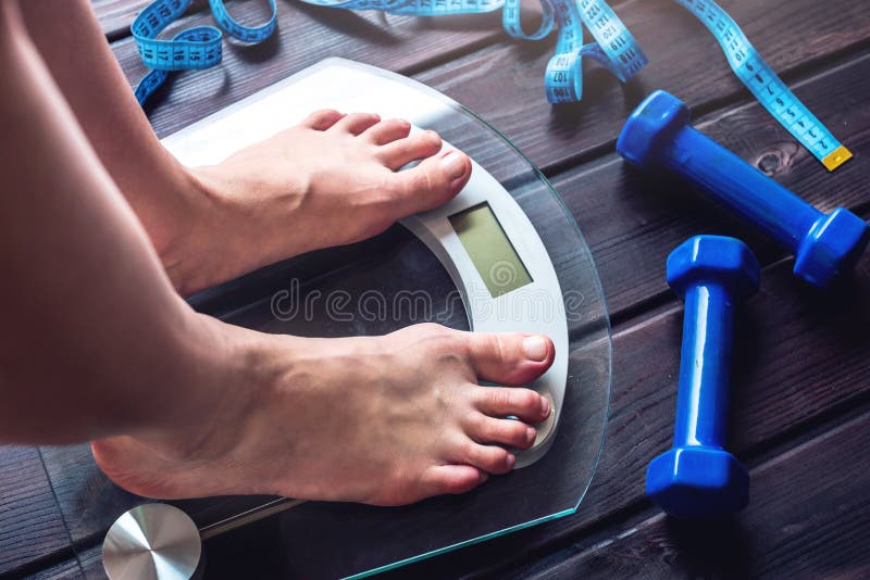 Female feet standing on electronic scales for weight control, dumbbells and measuring tape on wooden background. The concept of sports training, slimming and weight loss. Female feet standing on electronic scales for weight control, dumbbells and measuring tape on wooden background. The concept of sports training, slimming and weight loss