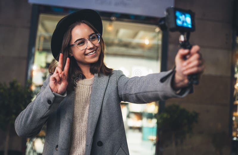Weibliche vlogger Datensatz mit Digitalkameravertretungssieg oder Frieden. Lächeln Frau, die selfie Video auf heller Nachtstadt wi stockfotografie