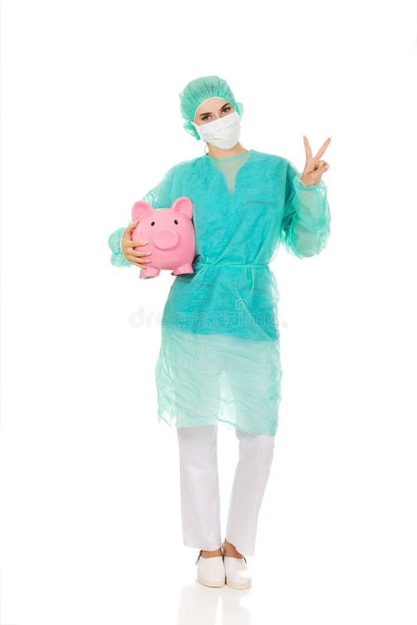 Weiblicher Chirurgdoktor mit Sparschwein zeigt Siegeszeichen lizenzfreie stockfotografie Weiblicher Chirurgdoktor mit Sparschwein zeigt Siegeszeichen lizenzfreie stockfotografie