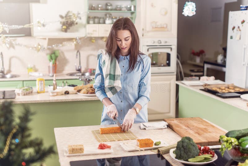 Female cook working in gloves making Japanese sushi rolls slicing them on bamboo mat standing in kitchen. Female cook working in gloves making Japanese sushi rolls slicing them on bamboo mat standing in kitchen.